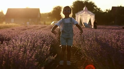 Adorable little girl is having fun and running against the background of a large lavender field on sunset. Hyperactive smiling little kid in sunglasses on nature. International Children's Day.