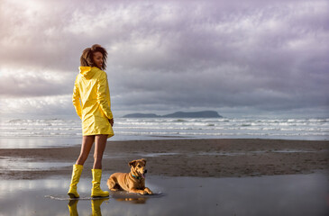 Young afro hairstyle black woman wearing yellow raincoat enjoying a winter relaxing walk at the beach with her dog. Generative AI.
