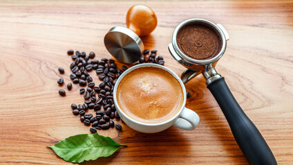 Top view of Espresso coffee cup and equipment of barista coffee tool portafilter with tamper and dark roasted coffee beans with green coffee leaf  on wooden table