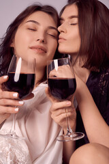 Valentine's day concept. Close-up portrait of two lesbian girls in satin negligee holding glasses of red wine, intimate moments of private life.