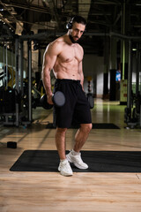 Fototapeta premium A brutal muscular man with a beard and a perfect body wearing headphones does exercises with dumbbells in the gym. Fitness, beauty