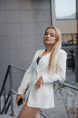 Blond stylish woman at oversized white jacket posing at the railing and steps on the city background