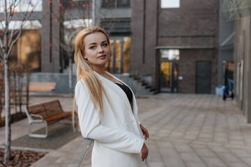 Fototapeta premium Blond stylish woman at oversized white jacket posing at the railing and steps on the city background