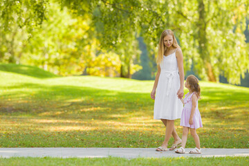 Little daughter and young adult mother in dresses speaking and walking on sidewalk at city park. Spending time together in beautiful warm sunny summer day. Side view.