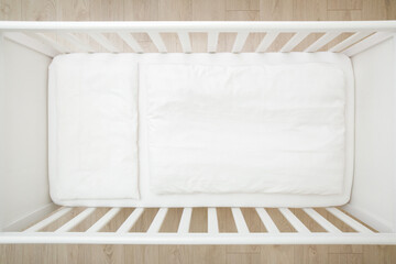 Empty white baby crib with mattress, sheet, pillow and blanket. Closeup. Top down view. - obrazy, fototapety, plakaty