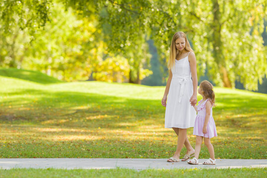 Little Daughter And Young Adult Mother In Dresses Speaking And Walking On Sidewalk At City Park. Spending Time Together In Beautiful Warm Sunny Summer Day. Side View.