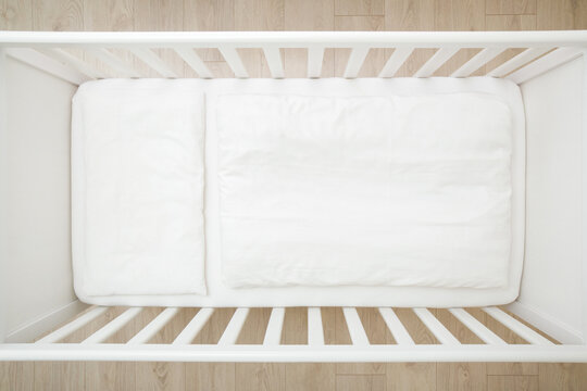 Empty white baby crib with mattress, sheet, pillow and blanket. Closeup. Top down view.