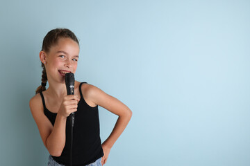 Portrait of a cute teenage girl singing, using microphone. Teenage girl in black t-shirt on an isolated blue background. Karaoke for kids, home entertainment for kids. Singer singing with microphone.