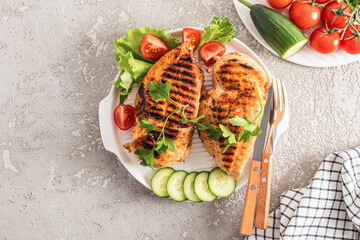 top view of the serving white plate with juicy grilled chicken fillet with fresh vegetables on a cement background. a copy of the space.