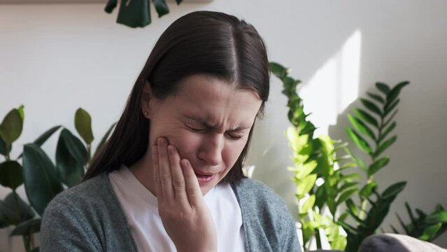 Close up of young woman feel terrible toothache after drink cold water. Brunette female sit on sofa at home touching cheek, feel hurt and suffering from sensitive tooth ache. Pain and cavities concept