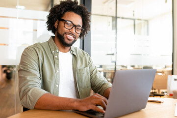 Portrait of smiling positive male office employee with computer indoors, African-American guy using...