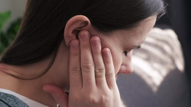 Close up of upset young brunette female holding painful ear, suddenly feeling strong ache. Unhealthy caucasian woman suffering from painful otitis sitting on sofa at home. Health problems concept