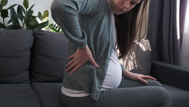 Side view of sad young pregnant woman suffers from back pain sitting on couch at home, future mother suffer from spine muscle ache backache. Concept pregnancy, motherhood, health and lifestyle