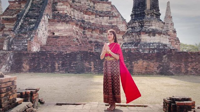 young woman dressed in traditional red Thai dress and golden ornaments stands sawasdee as a welcome and greeting in the ancient site Wat Chaiwatthanaram temple. Thai national costume
