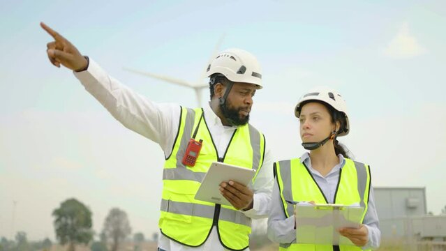 Front view of two african american man and woman in uniform discuss use laptop walking to verify that a wind turbine is running correctly and effectively, Renewable energy, technology, power, service