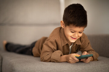Glad caucasian little child in casual watching video on smartphone, lies on sofa, enjoy spare time in living room