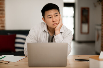 Upset bored middle aged asian man sitting in front of laptop at home, leaning on his hand and looking away