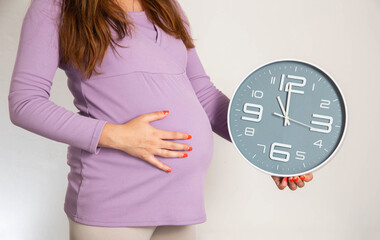 A pregnant girl in the 3rd trimester holds a wall clock against the background of a large belly....