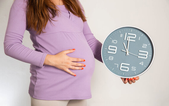 A pregnant girl in the 3rd trimester holds a wall clock against the background of a large belly. The concept of preterm birth, precursors and time to give birth.