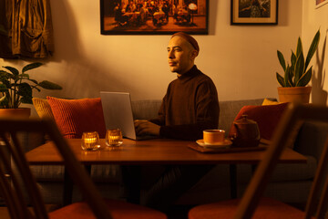 A caucasian man wearing a beanie and a knitted sweater sitting in a couch in a living room in a warm sunrise light writing on a laptop.