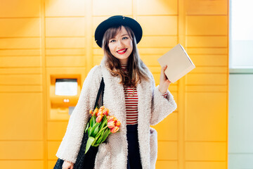 Portrait of smiling fashion woman with box near modern postal automatic mail terminal with self...