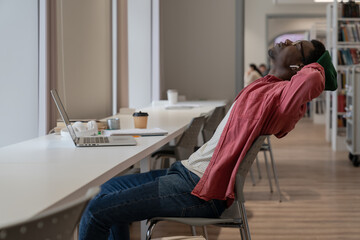 Exhausted young African American student guy feeling tired of e-learning, sitting at desk with laptop in library holding hands behind and resting from studying, taking break during online class