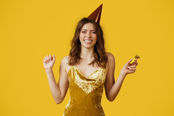 Excited lady in dress and birthday cap holding party horn, celebrating her special day on yellow...