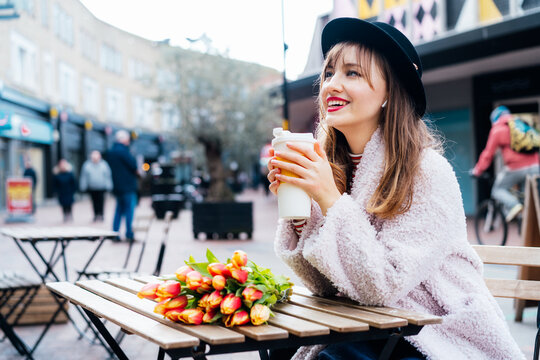 Stylish young smiling woman enjoying coffee from a reusable cup, listening to music, podcast using wireless earphones in a street cafe. Fresh tulips bouquet on the table. Springtime street fashion