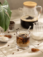 Coffee with milk in a glass with brown sugar,coffee beans and green monstera leaf on sunny tale table.Close up of hot drink.