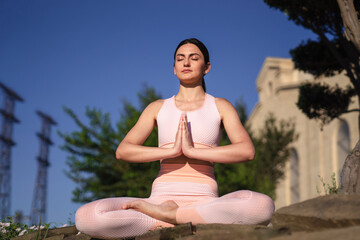 Fototapeta na wymiar Meditates in nature. A girl is engaged in yoga on the street, in a pink tracksuit does various exercises and yoga poses