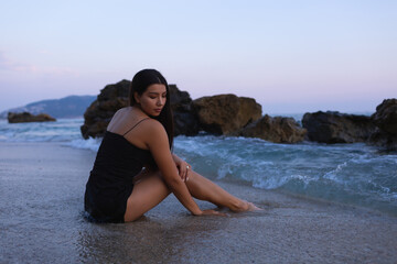 Fototapeta na wymiar A young girl in a short black dress is sitting on the sea coast. A beautiful woman is resting in the evening on a beautiful beach after sunset.
