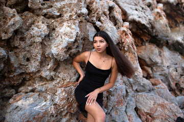 Beautiful brunette in a black dress posing near the rocks and the sea. Attractive brunette woman in a little black dress on the sea coast.