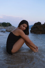 A young girl in a short black dress is sitting on the sea coast. A beautiful woman is resting in the evening on a beautiful beach after sunset.