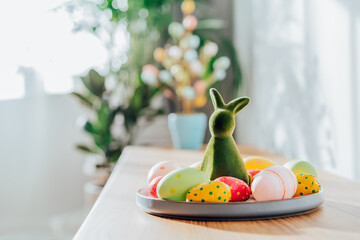 Easter home decor. Green bunny rabbit figurine and colored easter chocolate eggs on plate on wooden...