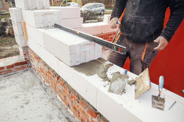 Builder installing masonry white blocks close up. Worker laying autoclaved aerated concrete blocks, working with adhesive and trowel. Process of house building at construction site