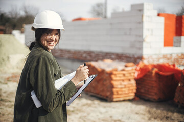 Young female engineer or construction worker in hardhat smiling against of building new modern house. Stylish happy woman architect with blueprints and plans at construction site