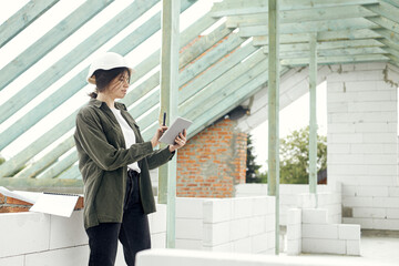 Young female architect with tablet checking blueprints against wooden roof framing of modern farmhouse. Stylish woman engineer in hard hat looking at digital plans at construction site