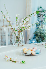 Blooming tree branches in the vase and colored easter eggs on the plate on white kitchen table with classic style interior background. Easter celebration at home. Festive spring vertical card.