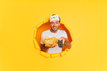 Horizontal shot of funny focused caucasian man holds bowl of chips and remote control chooses serial to watch has excited expression wears casual outfit and sleeping mask poses through torn paper hole