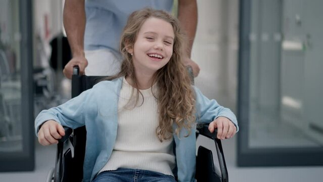 Young caregiver pushing little girl on wheelchair at hospital corridor, having fun together.
