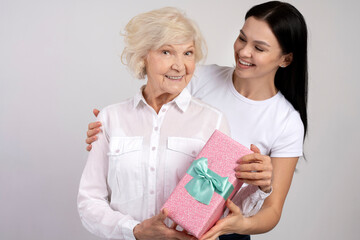 Lovely woman embracing her mom preparing gift case in pink package with bow, enjoying leisure time together isolated on white background. Mothers day concept