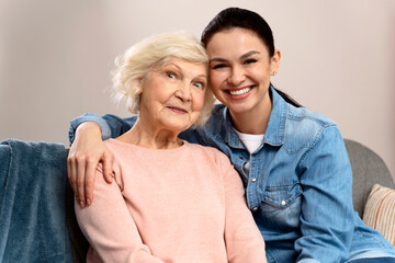 Portrait of old mother and daughter hugging at home. Happy senior mother and adult daughter embracing with love on sofa. Cheerful woman hugging from behind older mom and looking at the camera