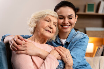 Cheerful woman hugging older mom with closed eyes. Happy senior mother and adult daughter embracing with love on sofa. Old mother and daughter hugging at home