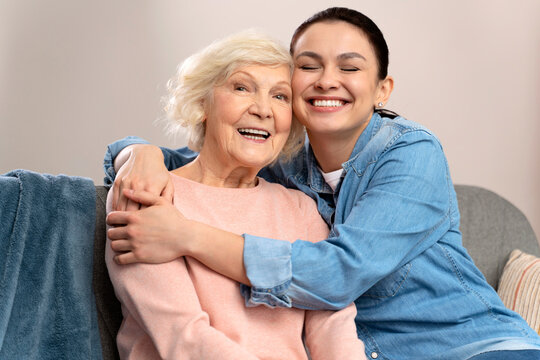 Happy Senior Mother And Adult Daughter Embracing With Love On Sofa. Old Mother And Daughter Hugging At Home. Cheerful Woman Hugging Older Mom With Closed Eyes