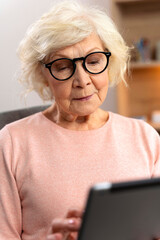 Vertical view of the smiling senior woman sitting with tablet on couch at home and hanging on...
