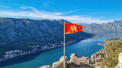 Montenegro flag blowing in wind with scenic view from St Ivan fortress on Kotor bay in sunny summer, Adriatic Mediterranean Sea, Montenegro, Balkan Peninsula, Europe. Fjord winding along coastal towns - obrazy, fototapety, plakaty