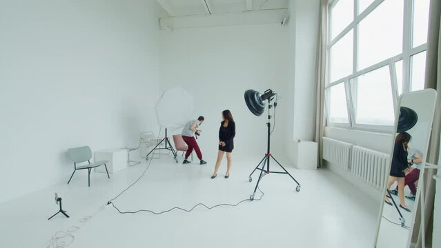 Male photographer shoots beauty portraits of female model in white photostudio. Camera man takes pictures of pretty girl, changes position of soft box. Young woman participates in photography session