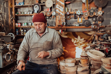 A senior man sitting in the workshop and processing wooden utensils in the old manual way
