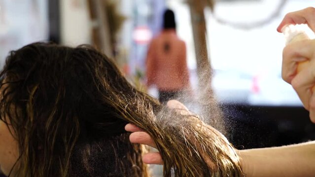 Hairdresser applies spray cosmetic product on young woman wet hair lock in professional beauty salon close view slow motion