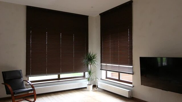 Wooden blinds closing down on large windows in the interior. Living room with armchair and houseplants near windows with wood blinds. Video of motorized jalousie in the smart house.